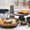 A styled tabletop arrangement of dark wood serving pieces on a light marble surface, including a large bowl with chips and dip, small bowls with olives and dip, a cake stand with two baked goods, glassware, a patterned vase, and a floral arrangement in the background.
