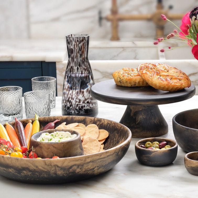 A styled tabletop arrangement of dark wood serving pieces on a light marble surface, including a large bowl with chips and dip, small bowls with olives and dip, a cake stand with two baked goods, glassware, a patterned vase, and a floral arrangement in the background.