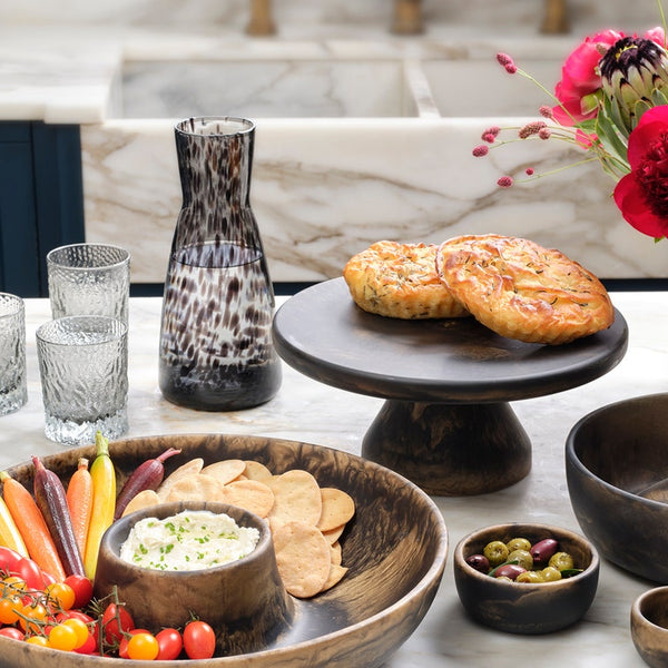 A styled tabletop food scene with wooden serving bowls, a dark pedestal plate holding two round baked goods, a patterned glass carafe, drinking glasses, olives, chips, dip, and a vase of red flowers on a marble surface.