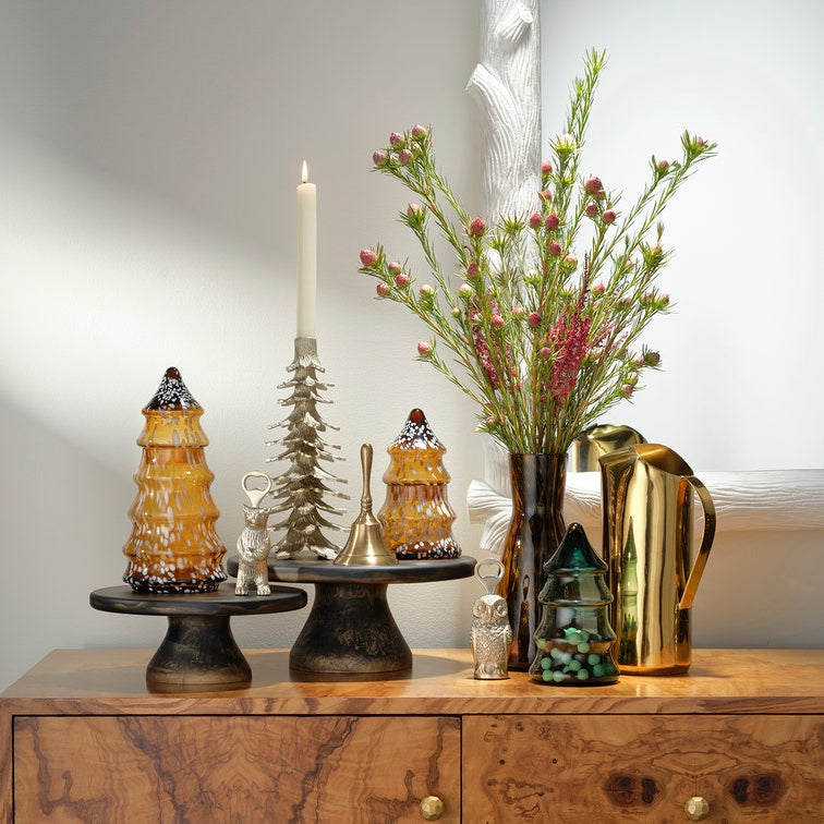 A styled holiday tabletop arrangement on a wooden cabinet against a light gray wall. The set includes amber glass Christmas tree ornaments with white snow details, a silver deer figurine, a silver bell, a silver Christmas tree, a white taper candle, a dark vase with pink and green floral stems, and a shiny gold pitcher.
