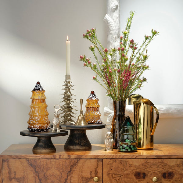 A styled holiday tabletop arrangement on a wooden cabinet against a light gray wall. The set includes amber glass Christmas tree ornaments with white snow details, a silver deer figurine, a silver bell, a silver Christmas tree, a white taper candle, a dark vase with pink and green floral stems, and a shiny gold pitcher.