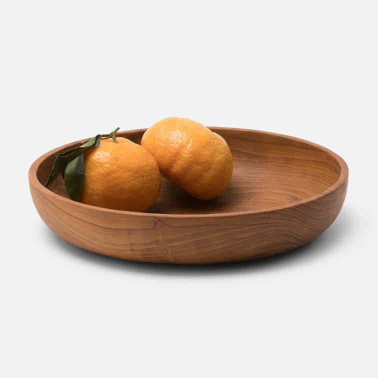 A round wooden bowl with a warm medium-brown finish, holding two oranges with green leaves, set against a clean light gray background.