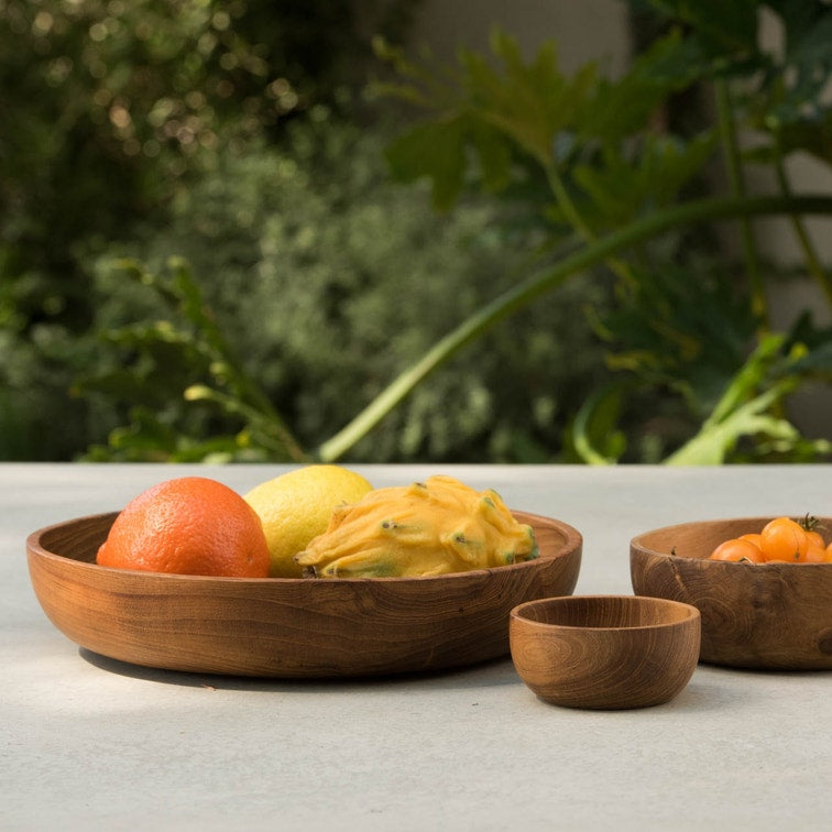 Three round wooden bowls of varying sizes on a light gray surface, styled with fruits including an orange, a yellow fruit, and small orange berries, with a blurred green garden background.