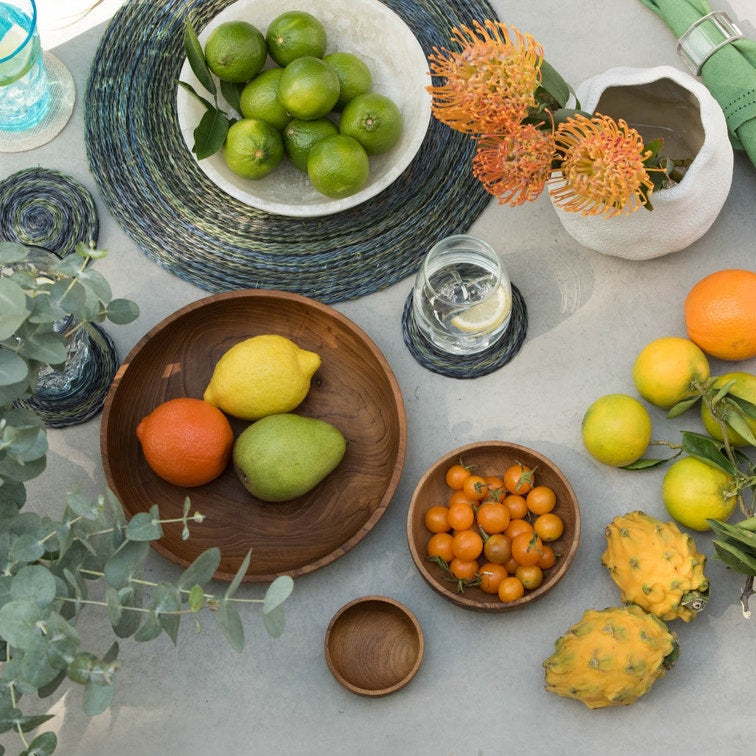 A styled flat-lay of citrus fruits, limes, dragon fruit, small yellow berries, green leaves, woven placemats, wooden bowls, a white vase with orange flowers, and a folded green cloth on a light gray surface.