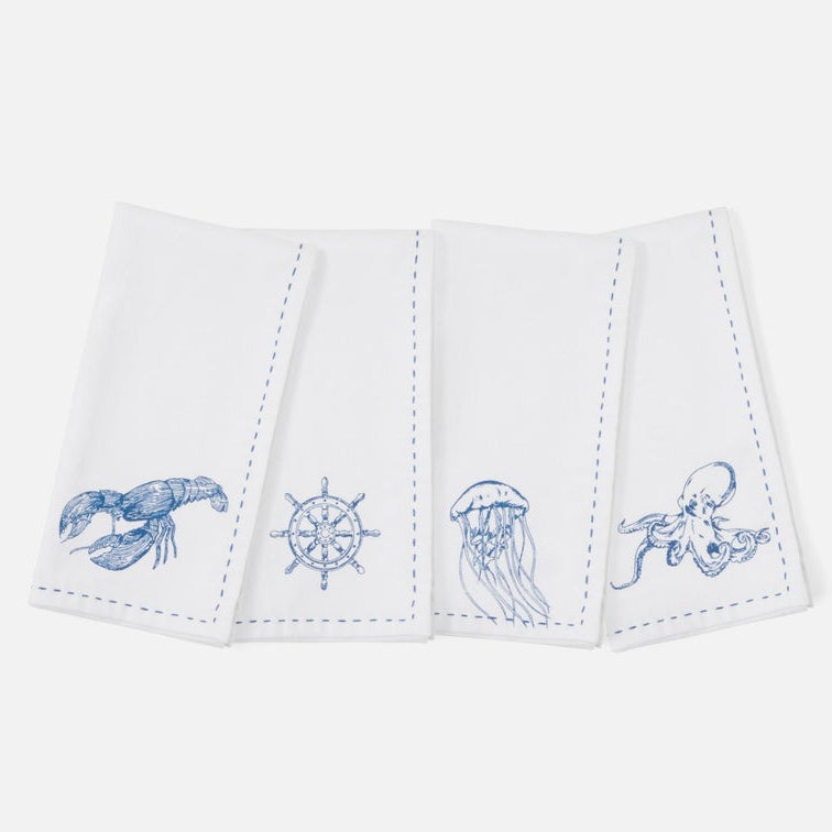 A set of four white napkins with blue embroidered sea life and nautical designs, including a lobster, ship's wheel, jellyfish, and octopus, arranged on a light gray background.
