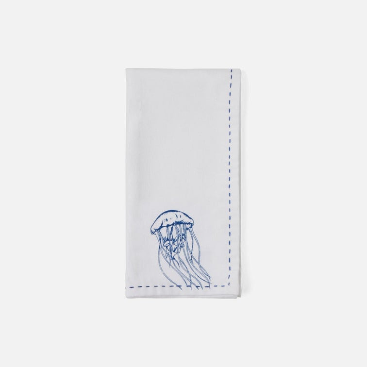 A white rectangular fabric napkin with a blue embroidered jellyfish design near the bottom and blue dashed stitching along the edges, shown on a light gray background.