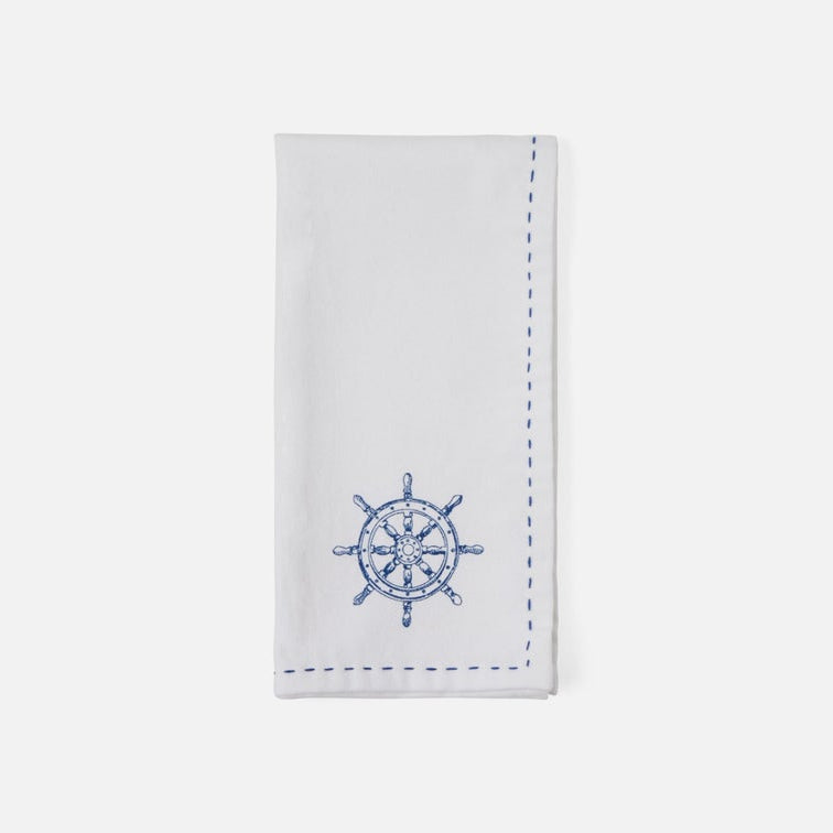 A white cotton napkin with blue embroidered details, featuring a ship's wheel motif near the bottom and dashed blue border stitching along the edges, displayed on a clean white background.