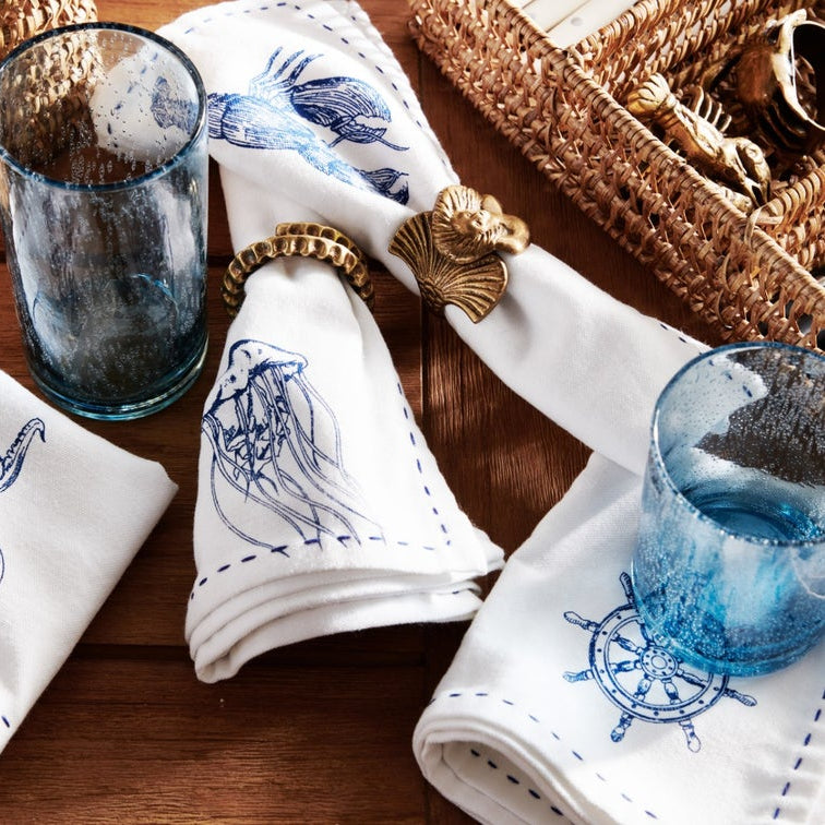 A styled tabletop scene with white napkins printed with blue nautical illustrations, including a jellyfish, octopus, ship wheel, and fish, arranged with blue-tinted drinking glasses and woven basket decor on a wooden surface.