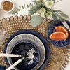 A styled tabletop setting featuring a navy blue speckled ceramic dinner plate with a matching bowl and a white-and-navy striped plate, accompanied by a smaller blue plate with orange slices and a knife. The place setting rests on a woven round placemat with metallic scalloped trim, with white flowers and greenery arranged above.