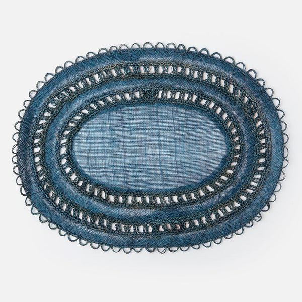 An oval blue woven placemat with intricate black and white embroidered borders, photographed from directly above on a clean white background.