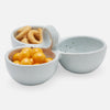 A three-compartment stoneware snack bowl in a pale blue-gray glaze, shown on a clean white background. The left compartment holds golden-brown ring-shaped snacks, the bottom-left compartment holds bright yellow round snacks, and the right compartment is empty.