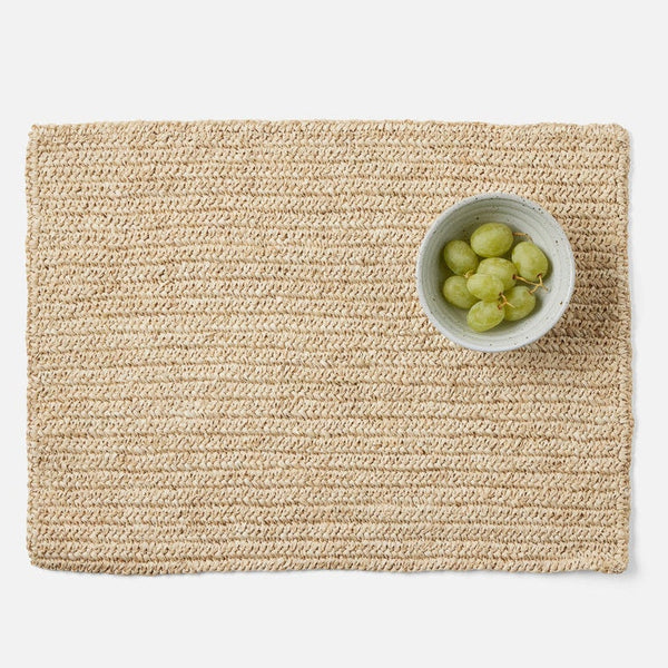 A rectangular woven placemat in a natural beige crochet texture, styled with a small white ceramic bowl of green grapes on a clean white background.