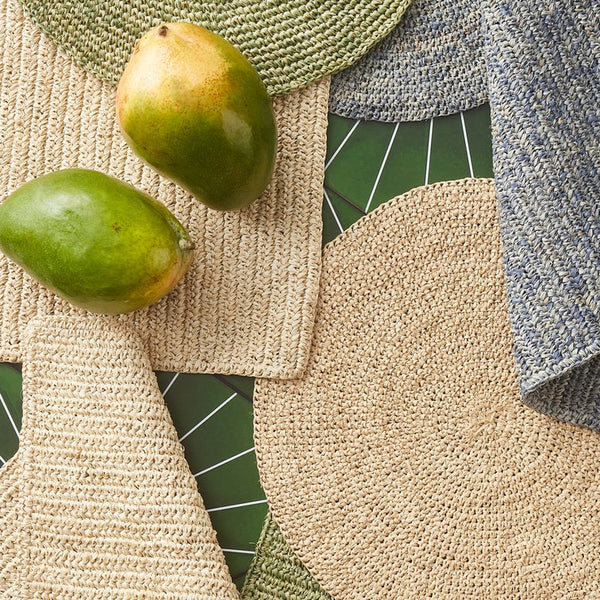 A styled flat-lay of woven home textiles, including a large round beige placemat, a blue-gray textured cloth, a green woven mat, and a beige rectangular mat, arranged on a dark green leaf-patterned surface with two green mangoes.