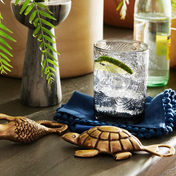 A textured clear drinking glass with a lime wedge, set on a blue crocheted coaster on a wooden table, with a turtle-shaped metal bottle opener and other rustic decor in the background.