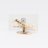 A small metallic gold bird-shaped place card holder with a textured finish, standing on a round base in front of a white card with a black 'B' logo.