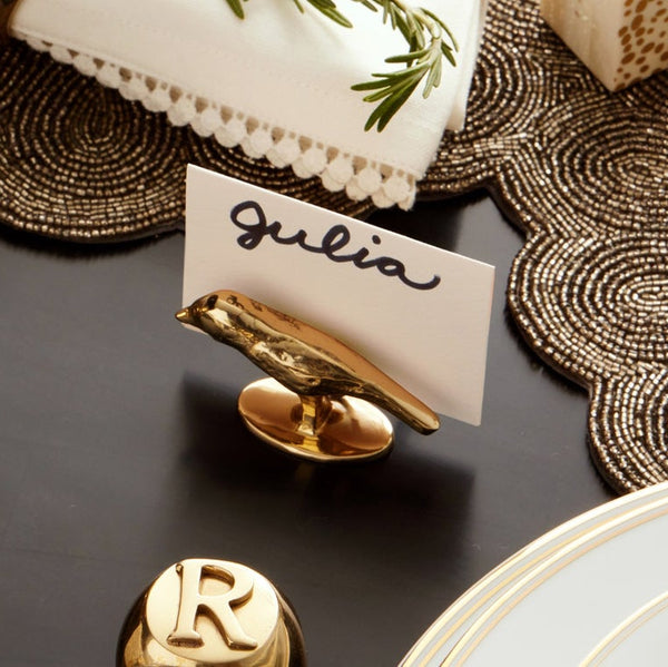 A gold bird-shaped place card holder on a dark table, holding a white card with the name 'Julia' in black script. The scene includes decorative placemats, a white plate, and other gold table accessories.