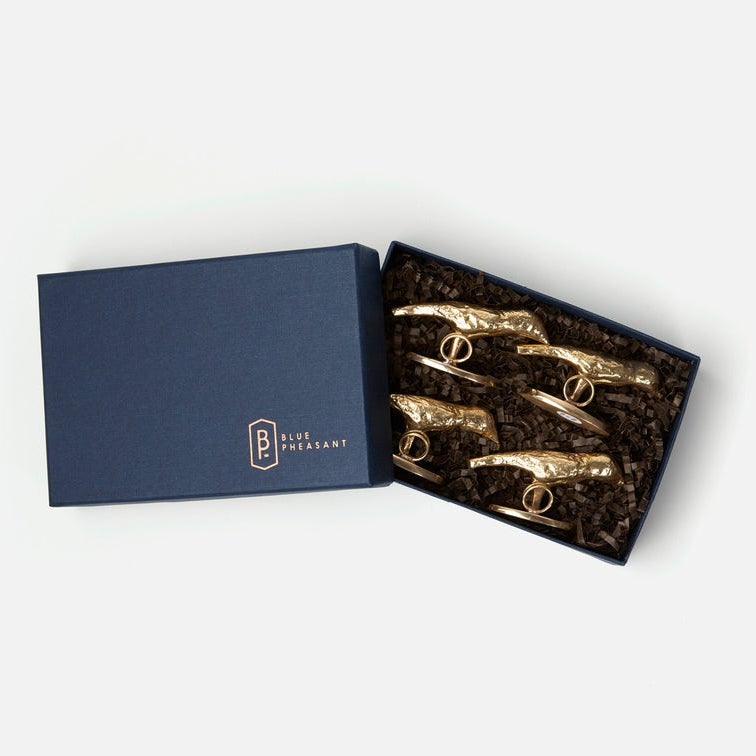 A navy blue gift box, partially open to reveal five gold-tone bird place card holders nestled in dark crinkle paper. The closed lid on the left features a gold logo that reads 'BLUE PLEASANT'.