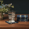 A set of indigo blue and cream ikat patterned coasters on a wooden table, with one coaster supporting a clear drinking glass of water and the other showing a stack of matching coasters.
