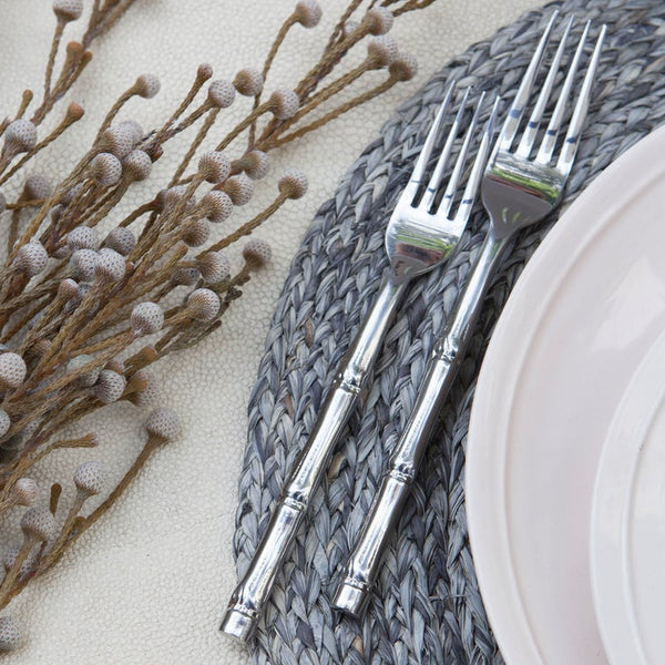 Two silver-toned metal forks with decorative handles, arranged on a gray woven placemat next to a white plate, with dried floral stems on a light beige textured surface.