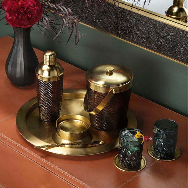 A luxury cocktail bar set featuring a hammered brass shaker, ice bucket, and matching glasses on a round brass tray, styled on a terracotta cabinet next to a black vase with red flowers.