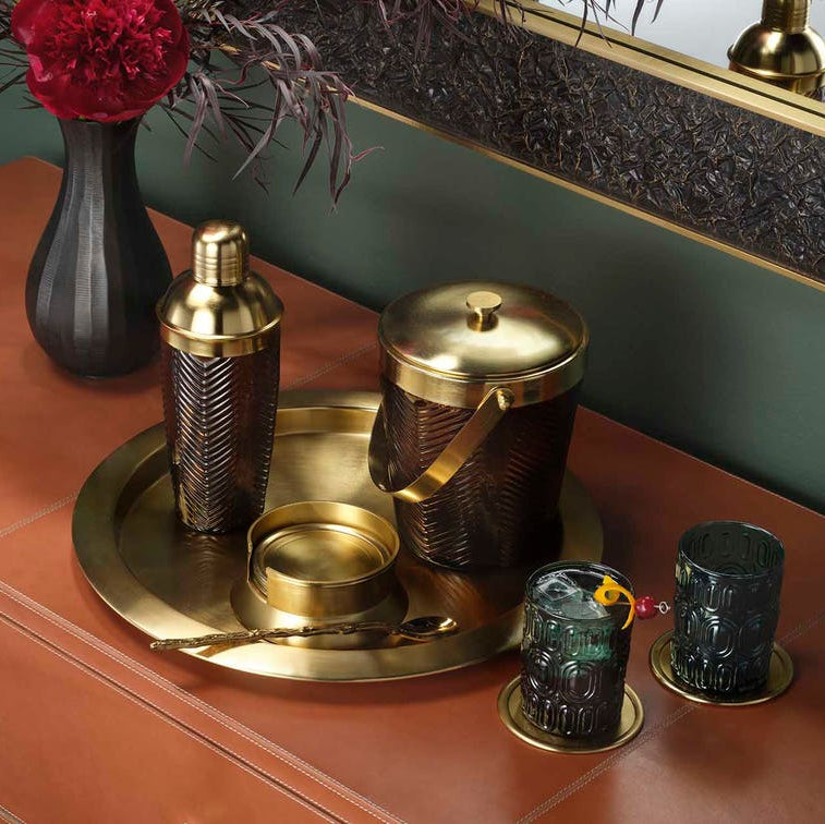 A luxury cocktail bar set featuring a hammered brass shaker, ice bucket, and matching glasses on a round brass tray, styled on a terracotta cabinet next to a black vase with red flowers.