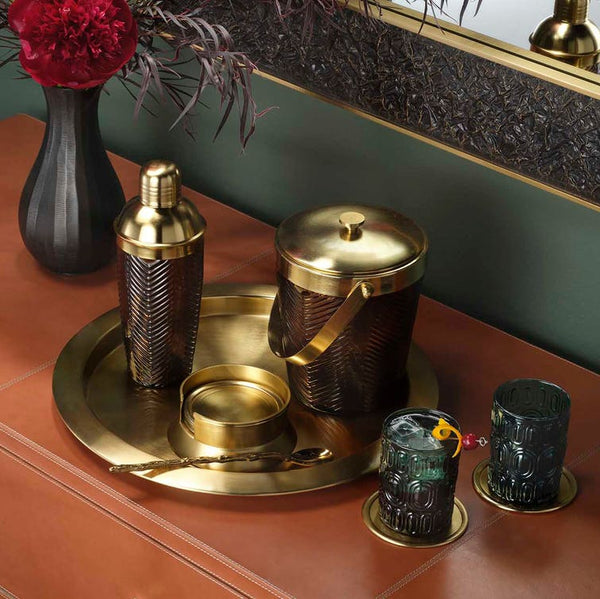 A luxury cocktail bar set featuring a hammered brass shaker, ice bucket, and matching glasses on a round brass tray, styled on a terracotta cabinet next to a black vase with red flowers.