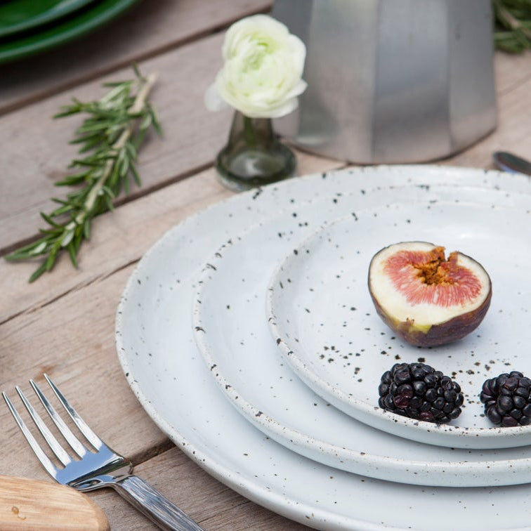 A styled tabletop setting with a stack of speckled white ceramic plates, a halved fig on the top plate, two blackberries, a fork, rosemary, and a small vase of white flowers on a wooden table.