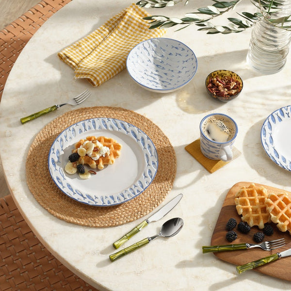 A styled breakfast table setting featuring a coordinated ceramic dinnerware set with blue hand-painted patterns on white, accompanied by green-handled cutlery, a yellow gingham napkin, and waffles on a wooden board.