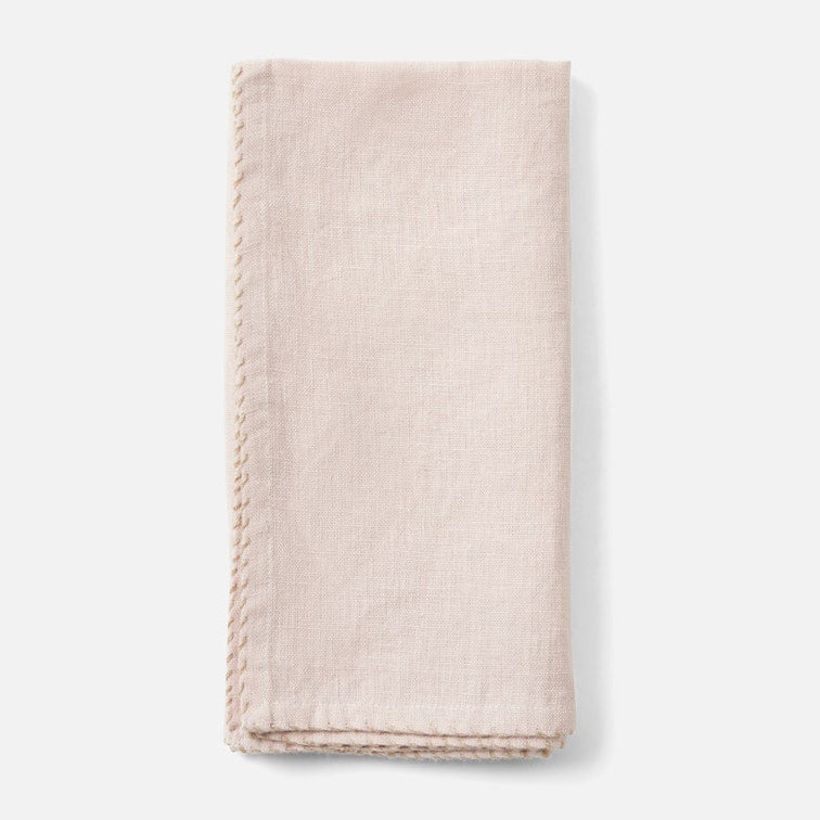 A folded light blush pink linen napkin with decorative hand-stitched borders, shown on a clean white background.