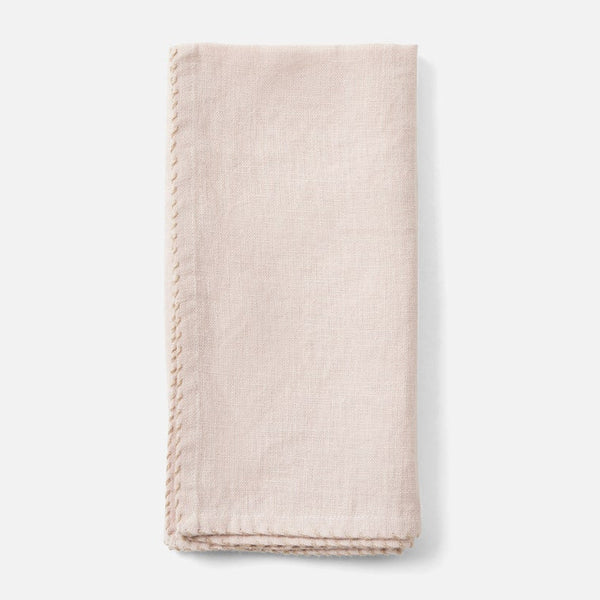 A folded light blush pink linen napkin with decorative hand-stitched borders, shown on a clean white background.