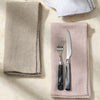A styled flat-lay of embroidered table linens on a light beige marble surface, featuring a beige linen napkin with braided edges, a blush pink linen napkin holding a silver knife and fork with black handles, and a folded white napkin with black embroidery at the top.