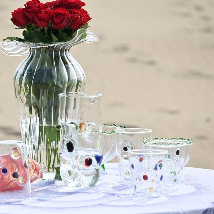 Clear glass pitcher and cups with colorful designs on a table with red roses in a vase.