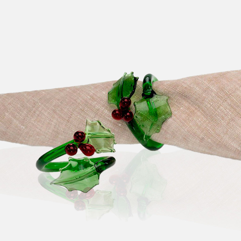 Green glass holly leaf and berry napkin rings on a beige napkin with a white background.