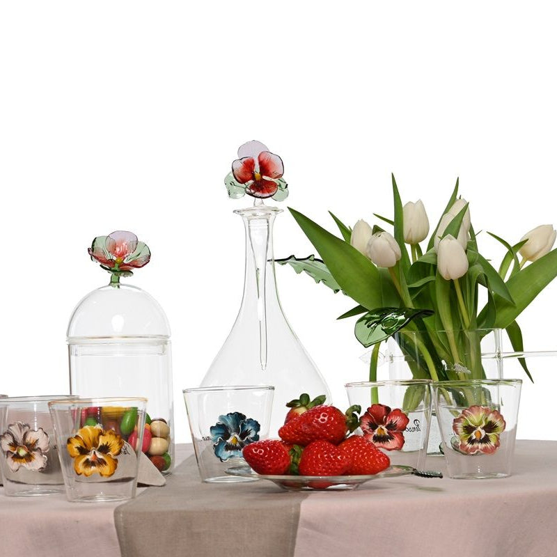 Decorative table setting with floral glasses, a decanter, and a vase of tulips on a white background.