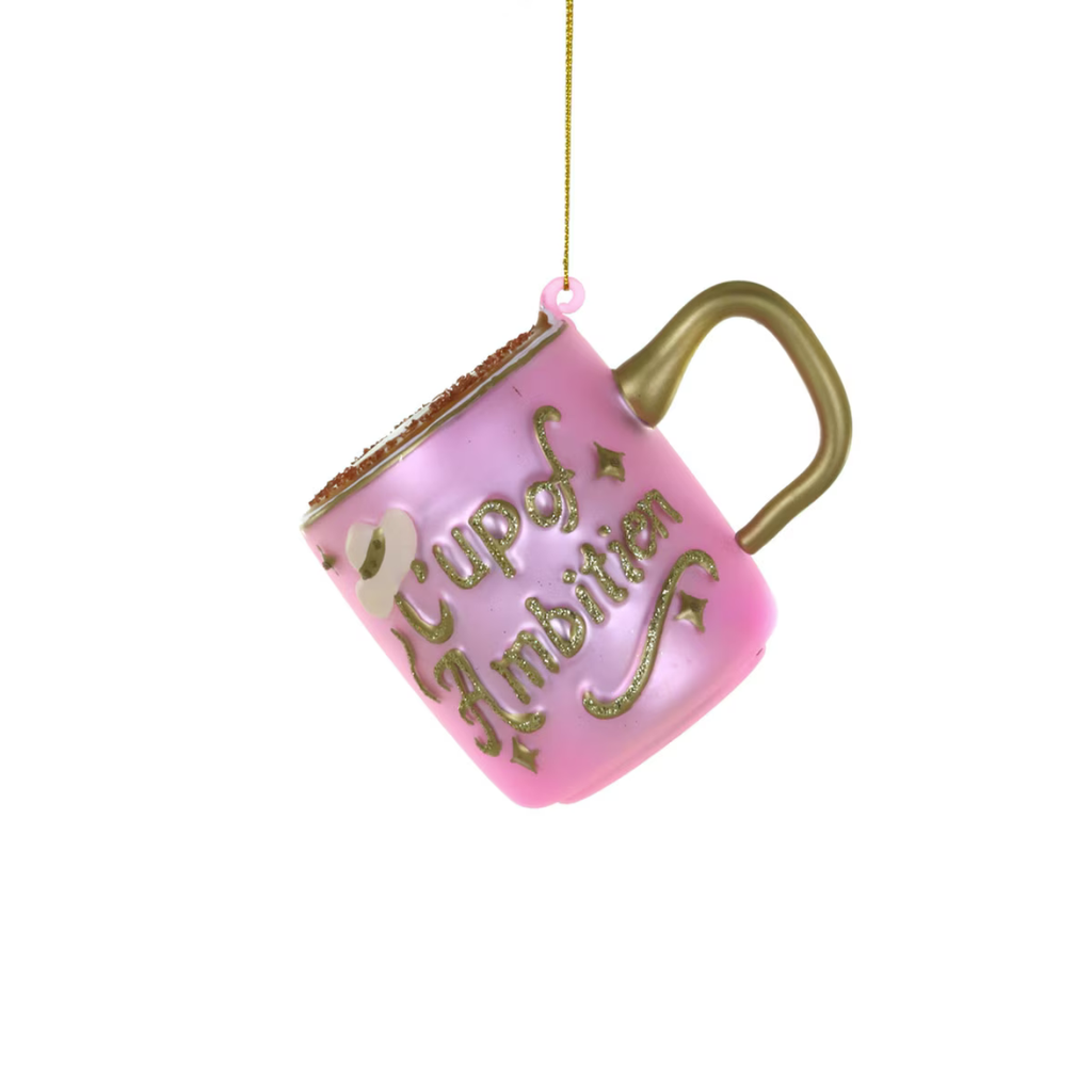 A pink Christmas ornament shaped like a coffee mug with the words 'Cup of Ambition' in gold glitter script, hanging by a gold string against a white background.