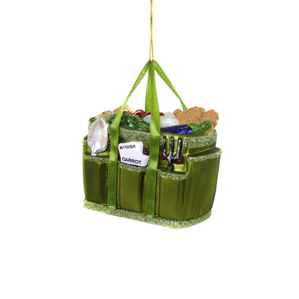 A green hanging ornament shaped like a garden tool basket with a gold hanging string, featuring a carrot, trowel, watering can, and other gardening-themed details on a white background.