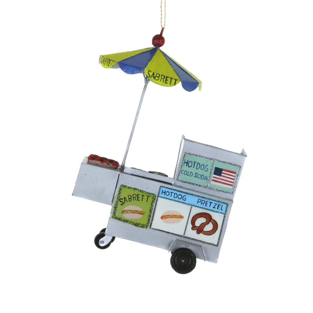 A hot dog cart Christmas ornament featuring a silver-colored metal cart with colorful painted signs, a blue and green umbrella, and black wheels, shown on a plain white background.
