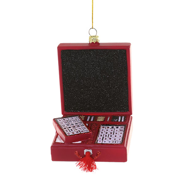 A red mahjong set ornament hanging from a gold string, with the box open to display a black textured back panel, a small red tile holder, two visible tile packs, and a red tassel at the front, on a plain white background.