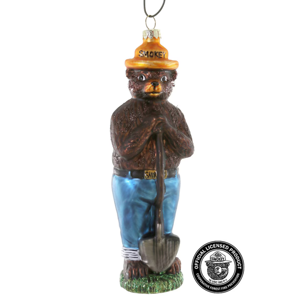 A glass Christmas ornament depicting a bear wearing a gold hat with the word 'SMOKEY' and blue pants, holding a shovel. A circular 'Official Licensed Product' seal is visible in the bottom right corner.