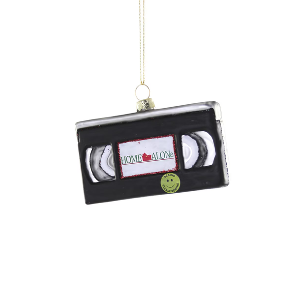 A VHS cassette-shaped Christmas ornament with a gold hanging string, featuring a black body, silver inner spools, a white label with red glittery text reading 'HOME ALONE', and a small green smiley-face sticker.