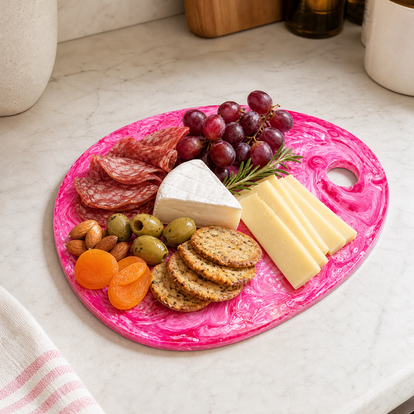A bright pink marbled charcuterie board with a handle, styled on a white marble kitchen counter with cheese, crackers, grapes, olives, almonds, and dried apricots.