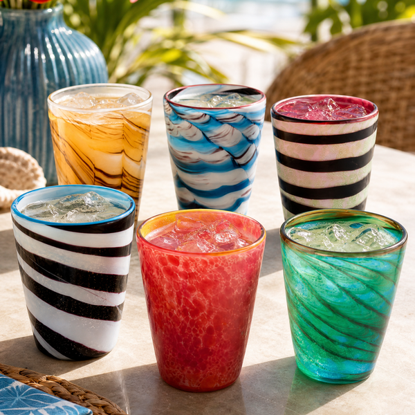Six colorful drinking glasses arranged on a stone table outdoors, with a blue vase, flowers, and wicker chair visible in the background. The glasses feature marbled, striped, and swirled patterns in black, white, blue, yellow, pink, red, and green.