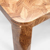 Close-up of a wooden table corner showing a polished geometric inlay pattern on the top and a matching wood grain leg, with a small brass doorknob visible on the front edge.