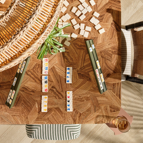 A top-down view of a wooden game table with rectangular tiles arranged in rows, alongside two dark green card stacks, a woven basket, and a striped chair in a styled indoor setting.