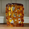 A sculptural, open-celled glass stool with an organic, cellular pattern in amber, gold, and deep reddish-brown tones, displayed on a wooden floor against a paneled wall.