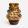 A sculptural, ribbed glass vase with a glossy amber-brown and black tortoiseshell pattern, set against a plain light gray background.