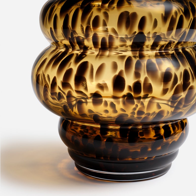 A close-up studio shot of a decorative glass vase with a rounded, tiered shape and a mottled amber, gold, and black pattern, set against a clean white background.