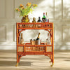 An orange-brown bamboo bar cart with gold-tone wheels and lattice-style side panels, styled with bottles, a flower vase, and an ice bucket against a cream paneled wall.