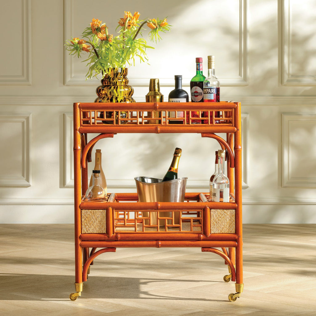 An orange-brown bamboo bar cart with gold-tone wheels and lattice-style side panels, styled with bottles, a flower vase, and an ice bucket against a cream paneled wall.