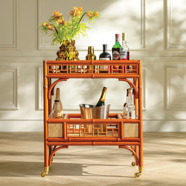 An orange-brown bamboo bar cart with gold-tone wheels and lattice-style side panels, styled with bottles, a flower vase, and an ice bucket against a cream paneled wall.